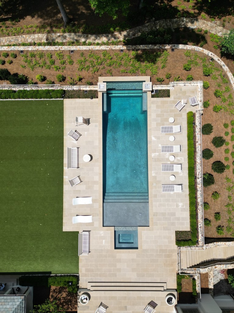 Aerial view of a modern backyard featuring a sleek swimming pool surrounded by stone tiles, sun loungers, and manicured landscaping.