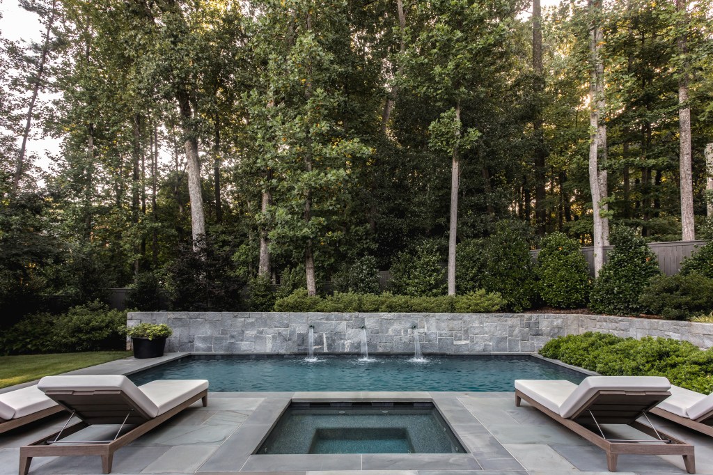 A tranquil outdoor space featuring a modern swimming pool surrounded by lounge chairs, set against a backdrop of tall trees and lush greenery.