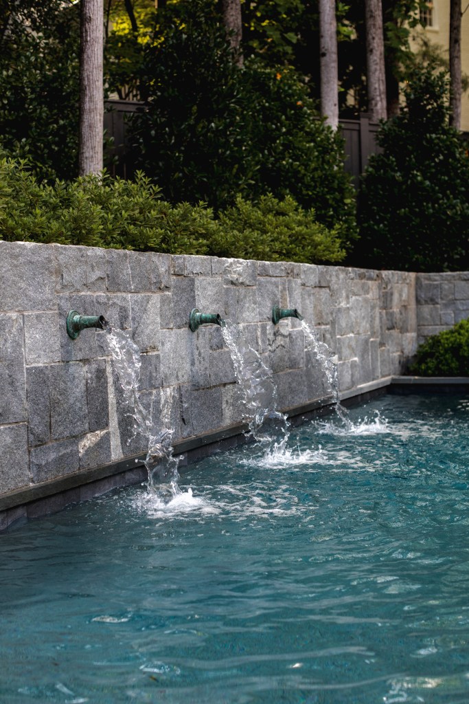 A stone wall with three water spouts flowing into a tranquil swimming pool surrounded by green foliage.