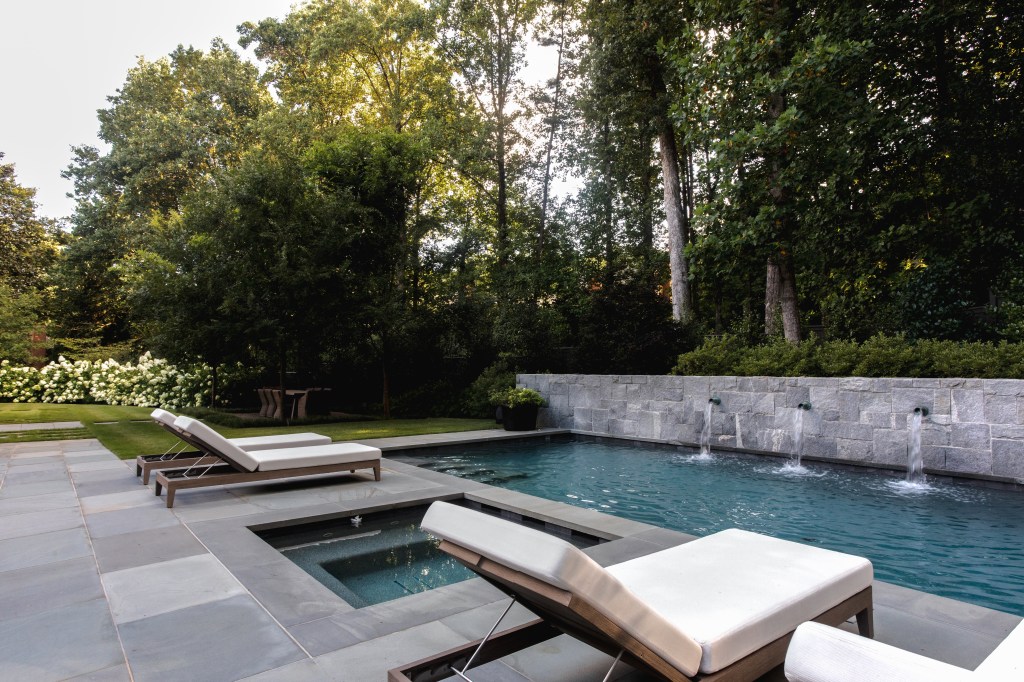 A serene backyard pool area featuring sun loungers, a small spa, and stone walls with water fountains, surrounded by lush greenery.
