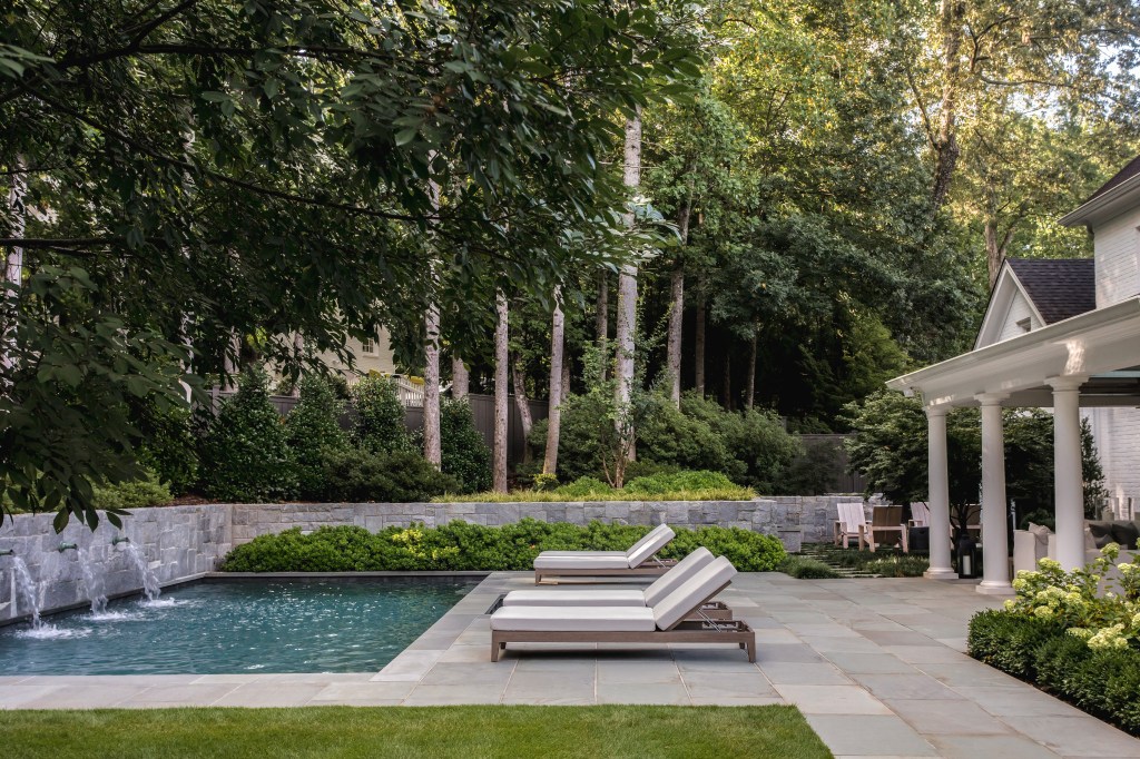 A tranquil backyard featuring a swimming pool with lounging chairs, surrounded by lush greenery and tall trees.