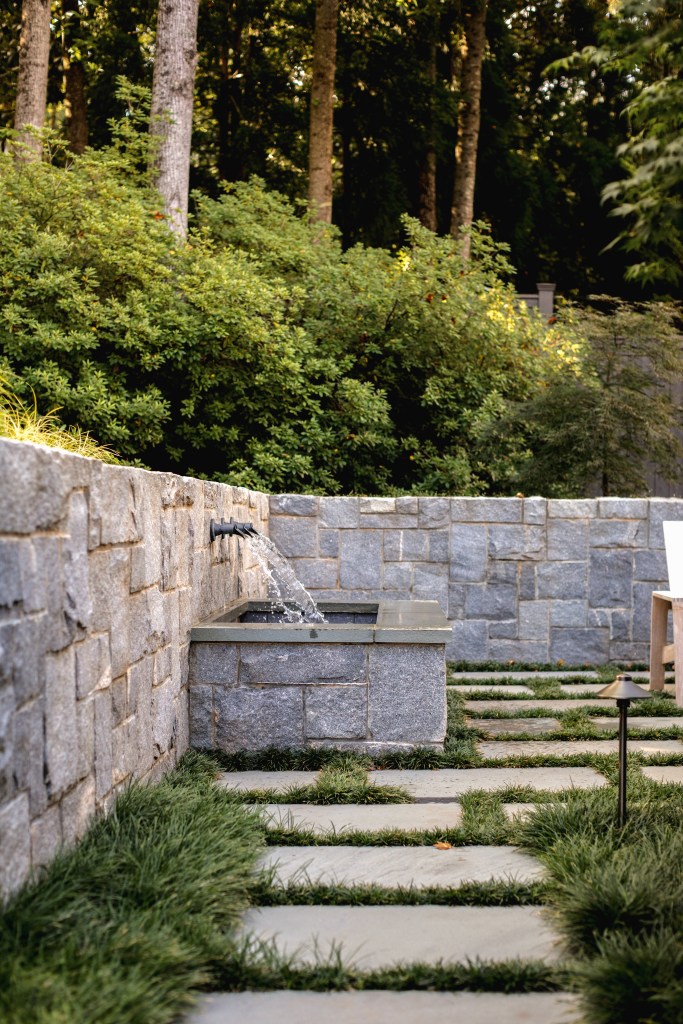 A stone water feature with flowing water, surrounded by grass paths and lush green foliage in a serene outdoor setting.
