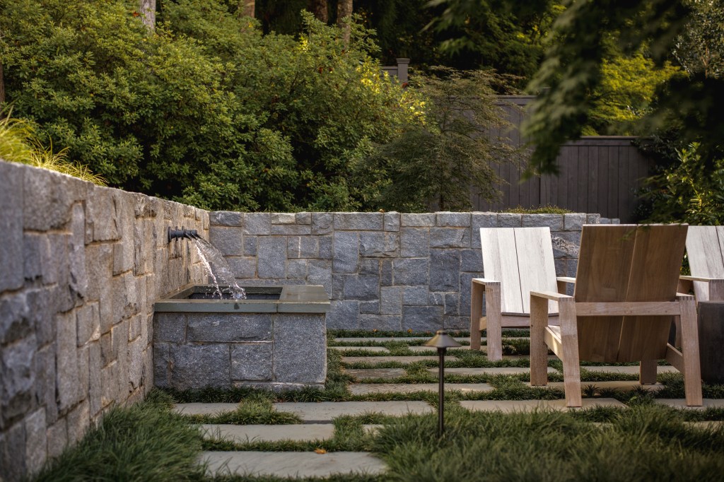 A serene outdoor space featuring a stone wall, a small water fountain, and two wooden chairs arranged on a stone pathway surrounded by lush greenery.