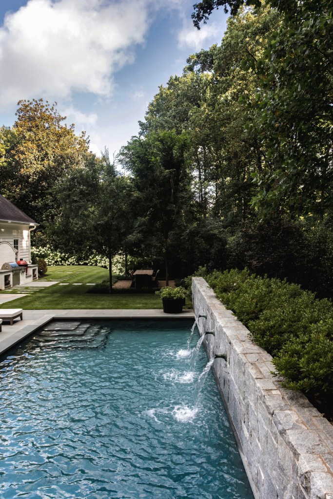 A serene backyard view featuring a swimming pool with a stone wall waterfall, surrounded by lush greenery and trees under a partly cloudy sky.