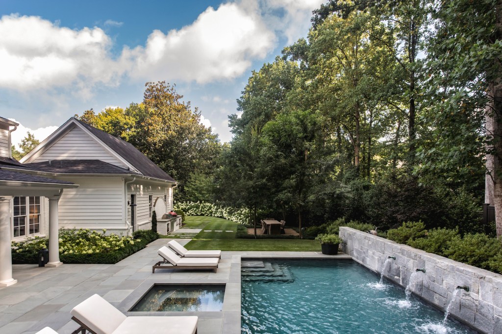 An elegant backyard featuring a swimming pool surrounded by lounge chairs, set against a backdrop of lush greenery and trees.