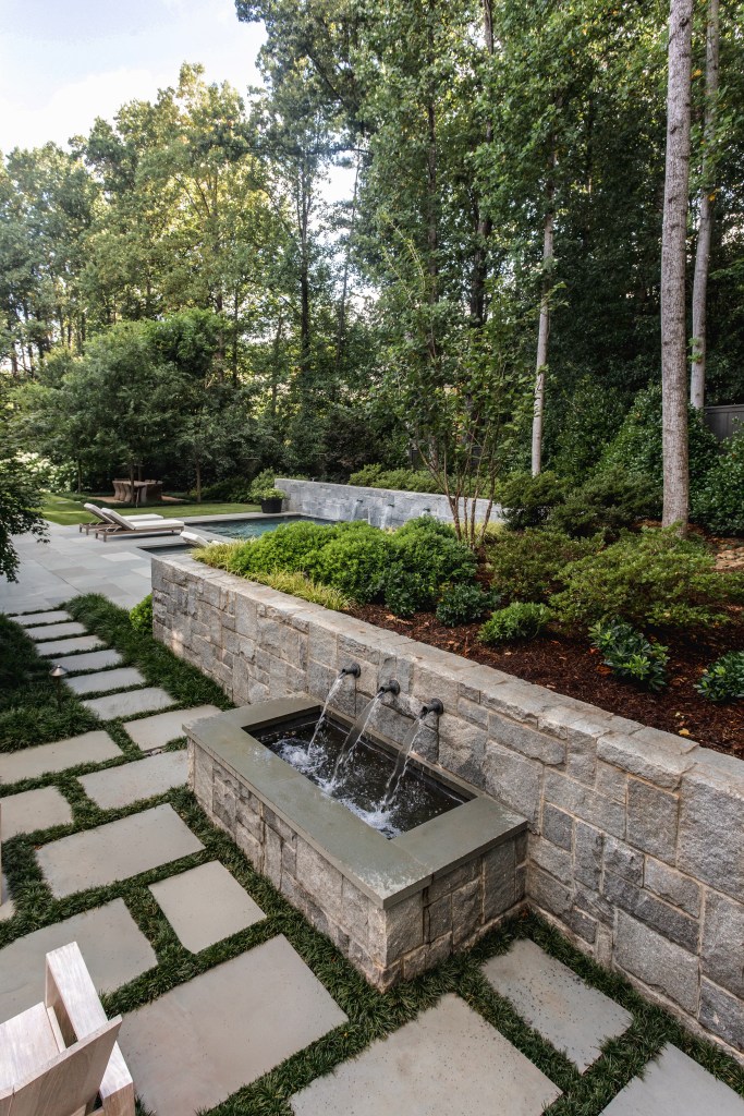 A serene outdoor landscape featuring a stone wall with three water spouts, surrounded by lush greenery and a pathway made of stone slabs.