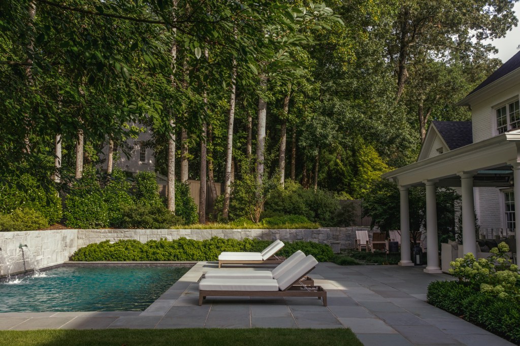 A serene outdoor patio with two white lounge chairs beside a swimming pool, surrounded by lush greenery and trees.