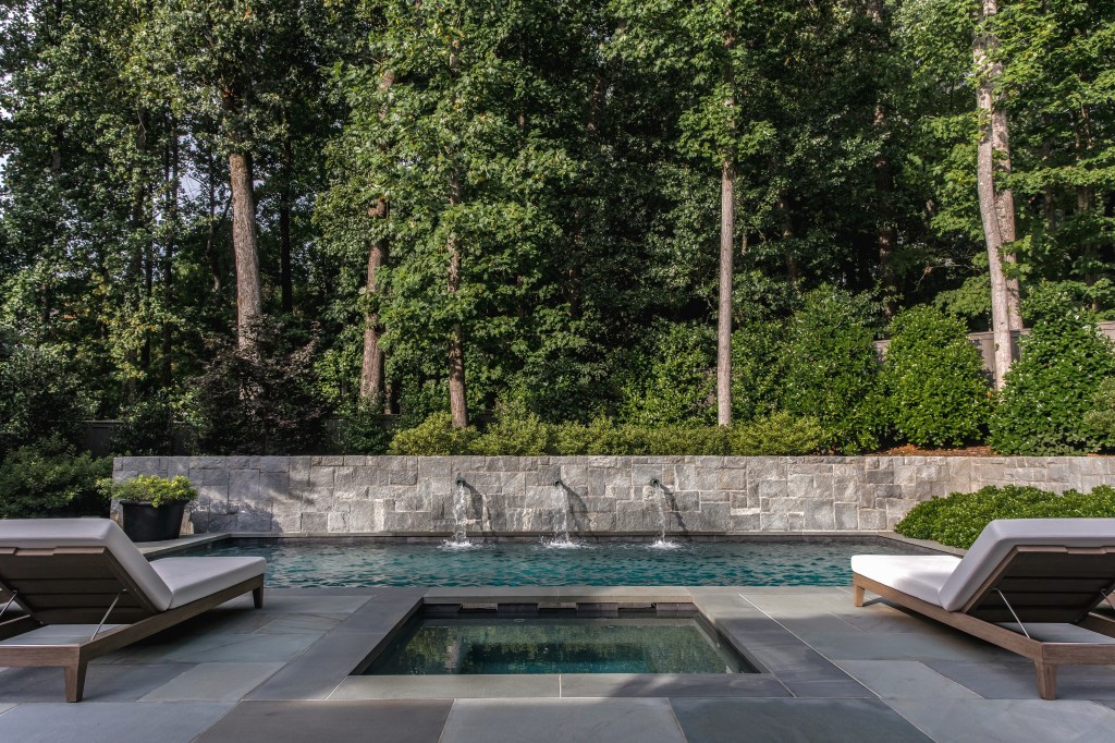 A serene backyard with a blue swimming pool featuring three water fountains, surrounded by lush green trees and shrubs. Lounge chairs are positioned on the patio made of stone tiles.