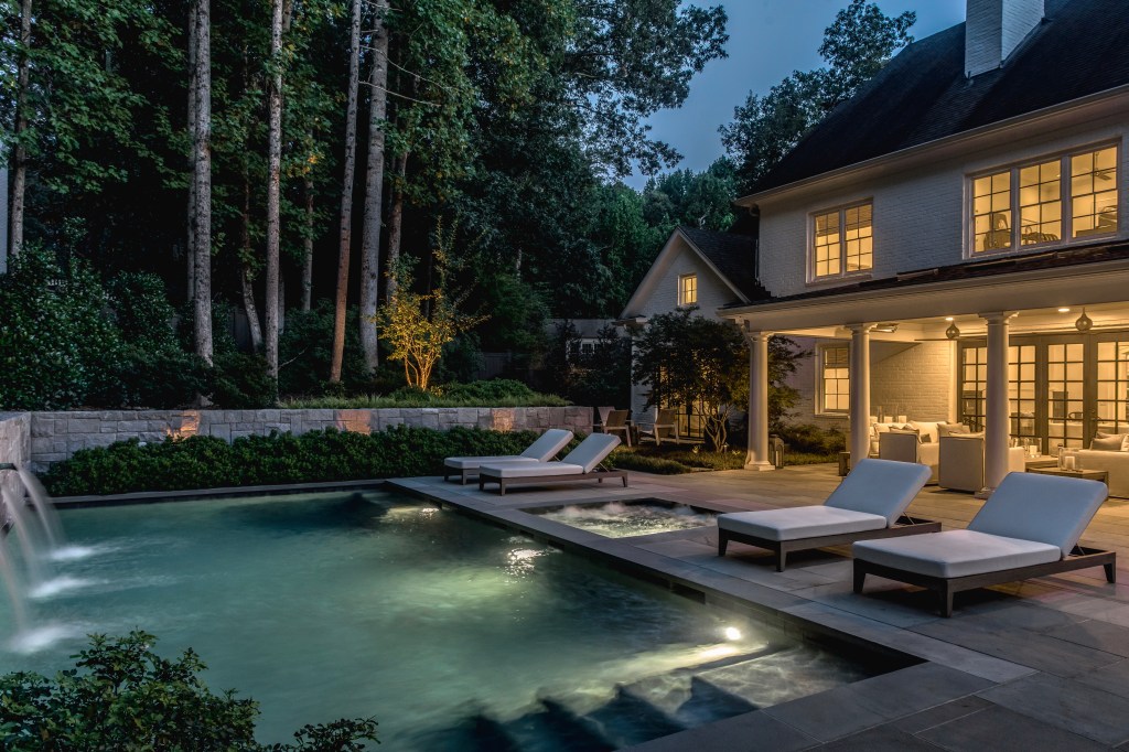 A tranquil backyard scene at night featuring a modern swimming pool with lounge chairs, surrounded by lush greenery and illuminated by soft lights. The house in the background has warm lights glowing through its windows.
