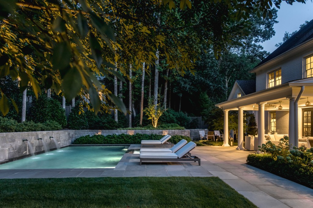 Nighttime view of a modern backyard featuring a swimming pool with illuminated water, surrounded by lounge chairs and a well-landscaped garden, adjacent to a cozy home.