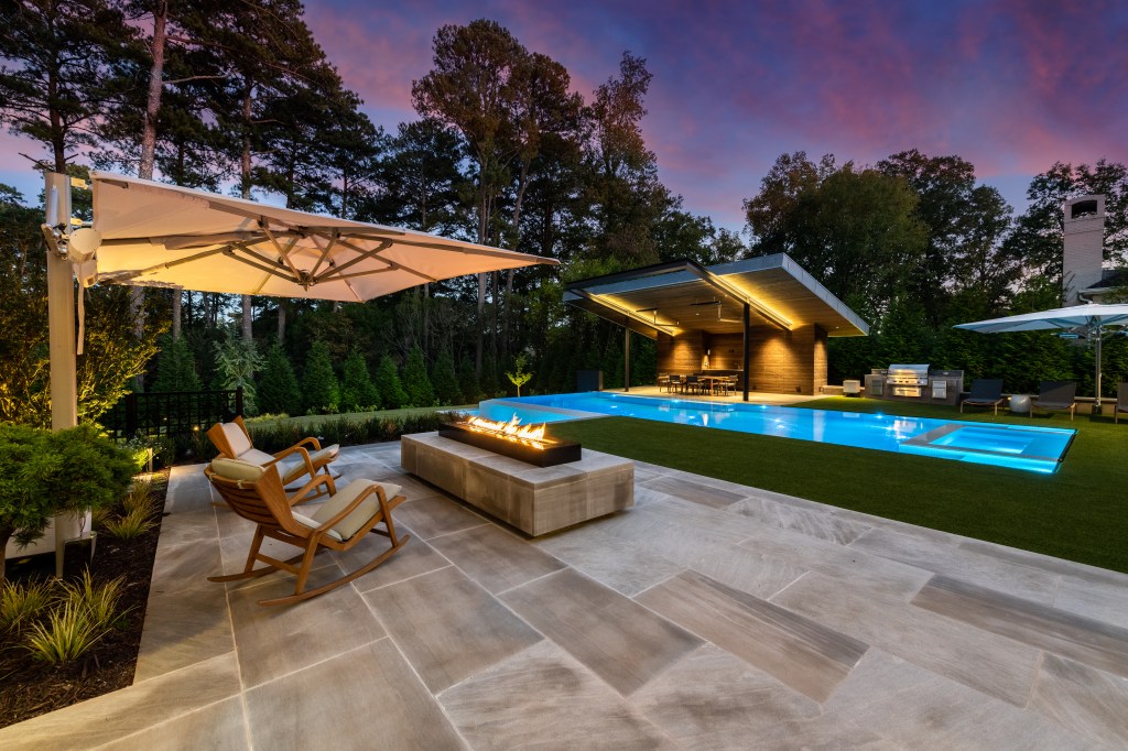 Modern backyard with a swimming pool, outdoor seating, and a dining area under a shelter, illuminated at dusk.