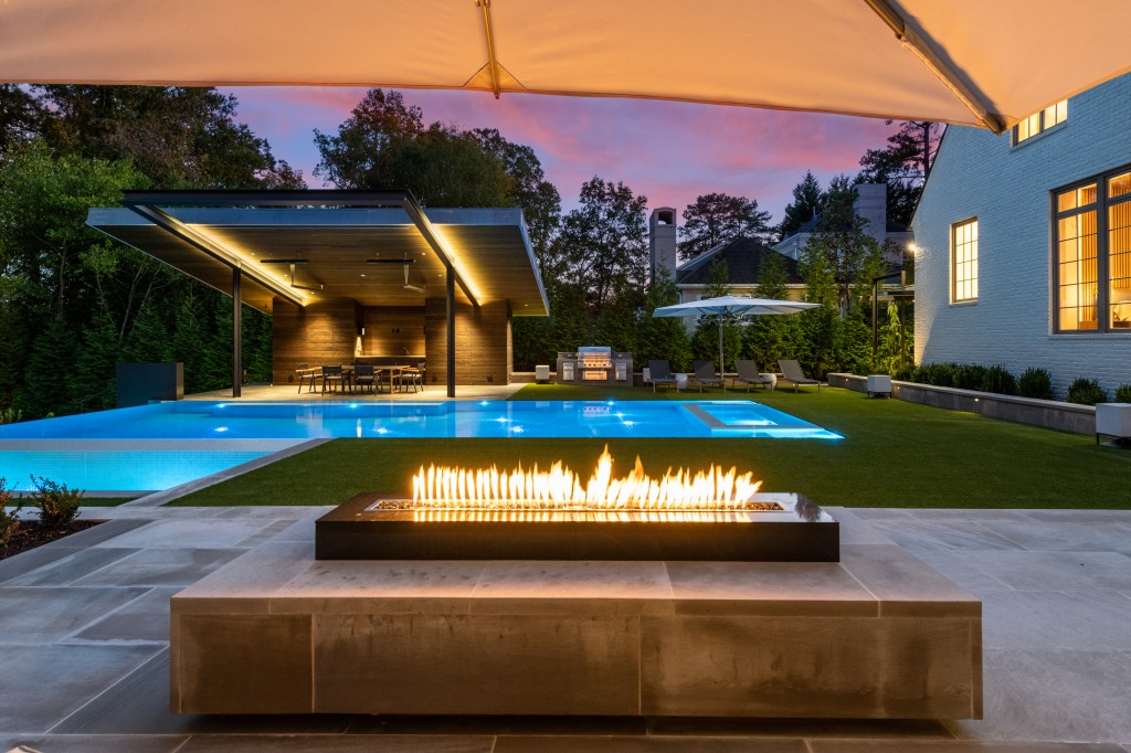 Modern outdoor living space featuring a pool, fire pit, and covered patio area with seating, illuminated at dusk.