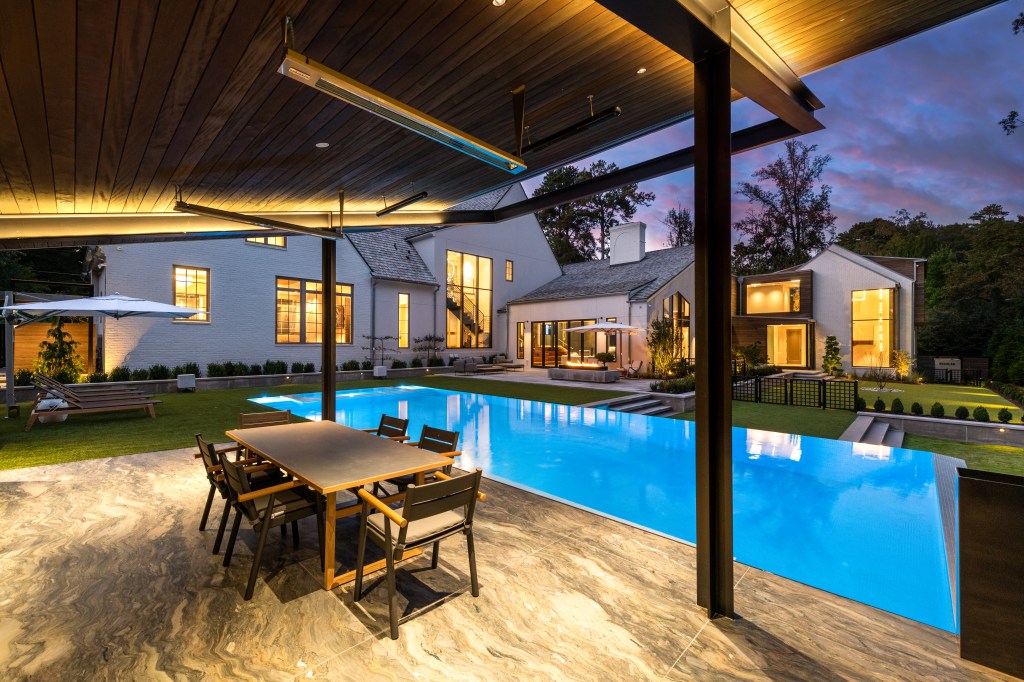 A modern home with a spacious backyard featuring a swimming pool, outdoor dining table, and lounge area, set against a colorful twilight sky.