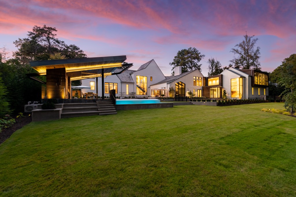 A modern house with large windows and a sleek design, featuring a swimming pool and an outdoor entertainment area, set against a colorful sunset sky.