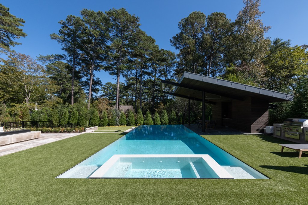 A modern backyard featuring a sleek swimming pool surrounded by lush greenery and trees, with a covered patio area and outdoor kitchen.