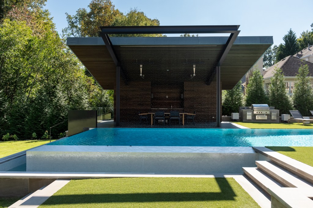 A modern outdoor pool with a sleek, rectangular design, surrounded by a well-maintained lawn and greenery. A stylish pavilion sits in the background, featuring a wooden structure with outdoor seating and a grill area.