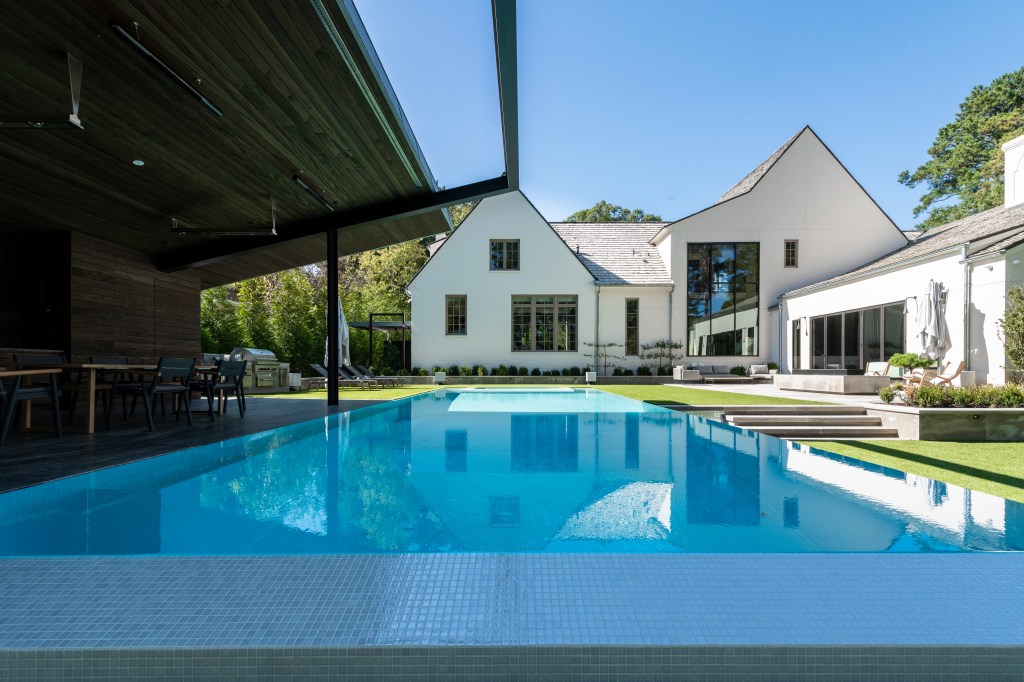 A modern home with a rectangular swimming pool, surrounded by green landscaping and outdoor seating areas, under a clear blue sky.