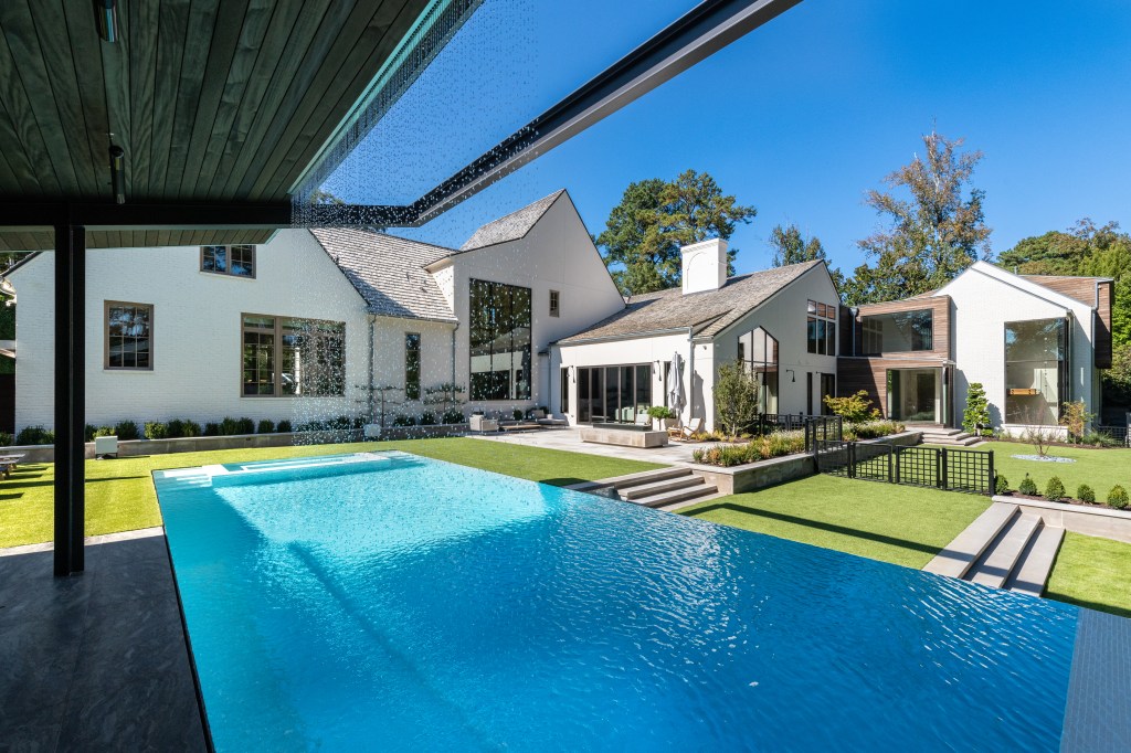 A modern backyard featuring a swimming pool with clear blue water, surrounded by lush green grass and well-maintained landscaping. In the background, several contemporary-style houses are visible under a bright blue sky.