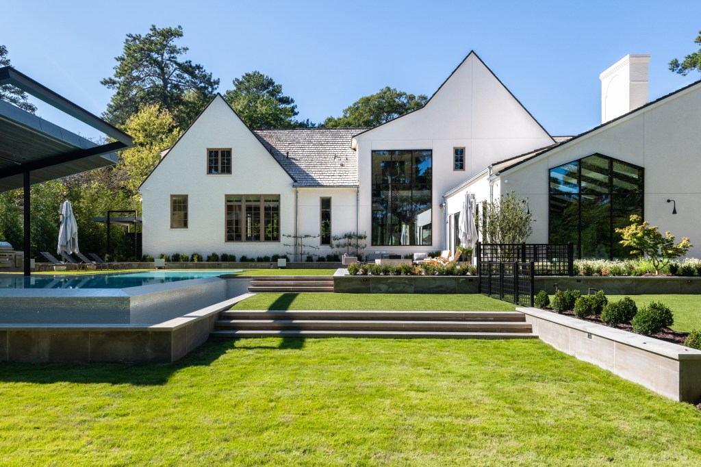 Modern white house with large windows, surrounded by green lawn and landscaped garden, featuring a swimming pool and steps leading to the backyard.